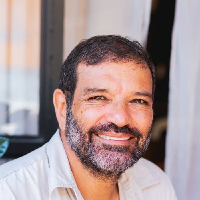 A man with a greying beard smiles, standing beside a window
