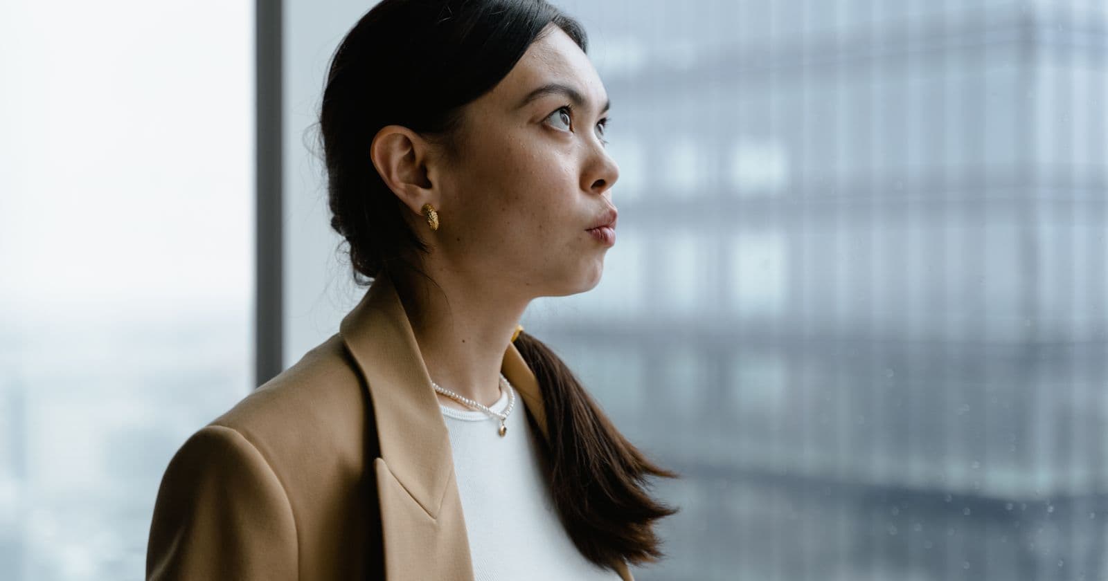 A woman in a camel blazer looks thoughtfully out of an office window