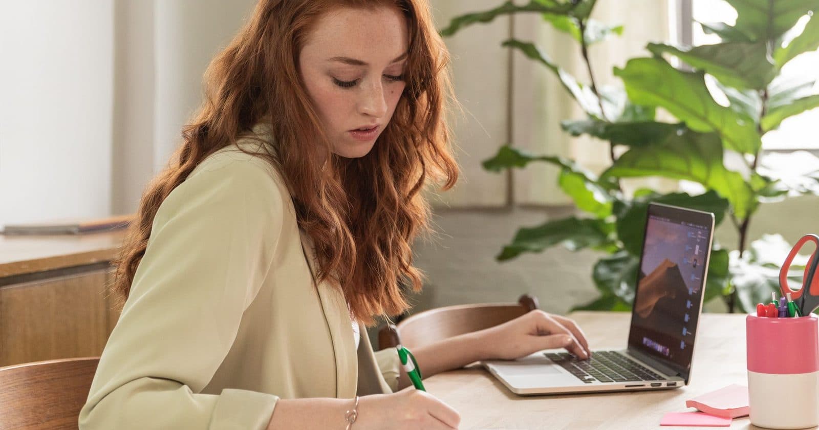 A woman at a wooden table by a window writes a note by hand, one hand on her laptop keyboard