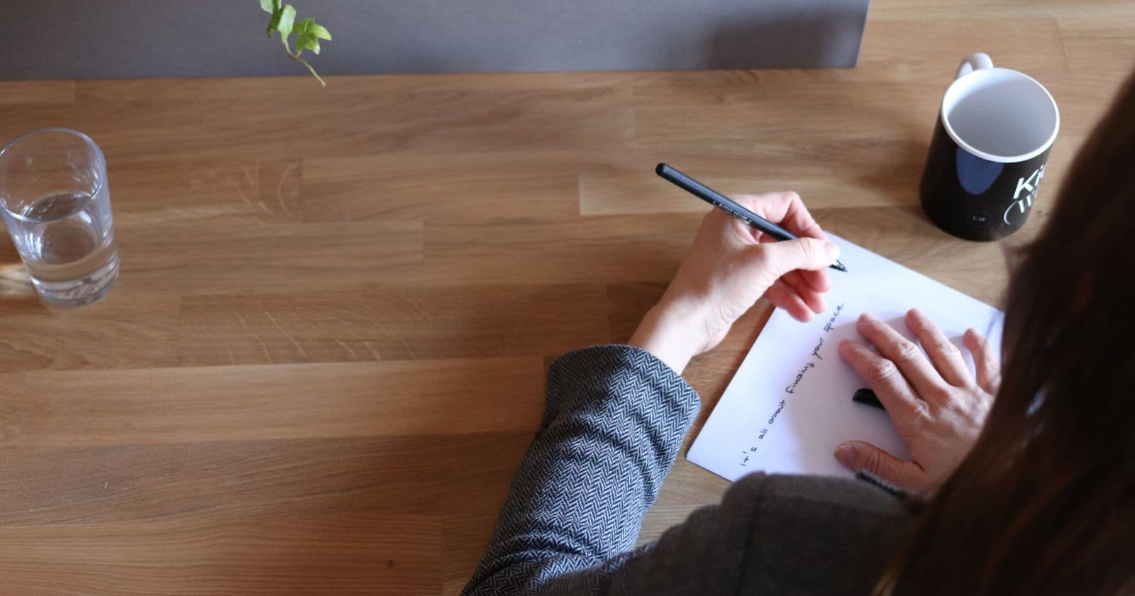 A woman writes notes on paper at a wooden desk, seen from above