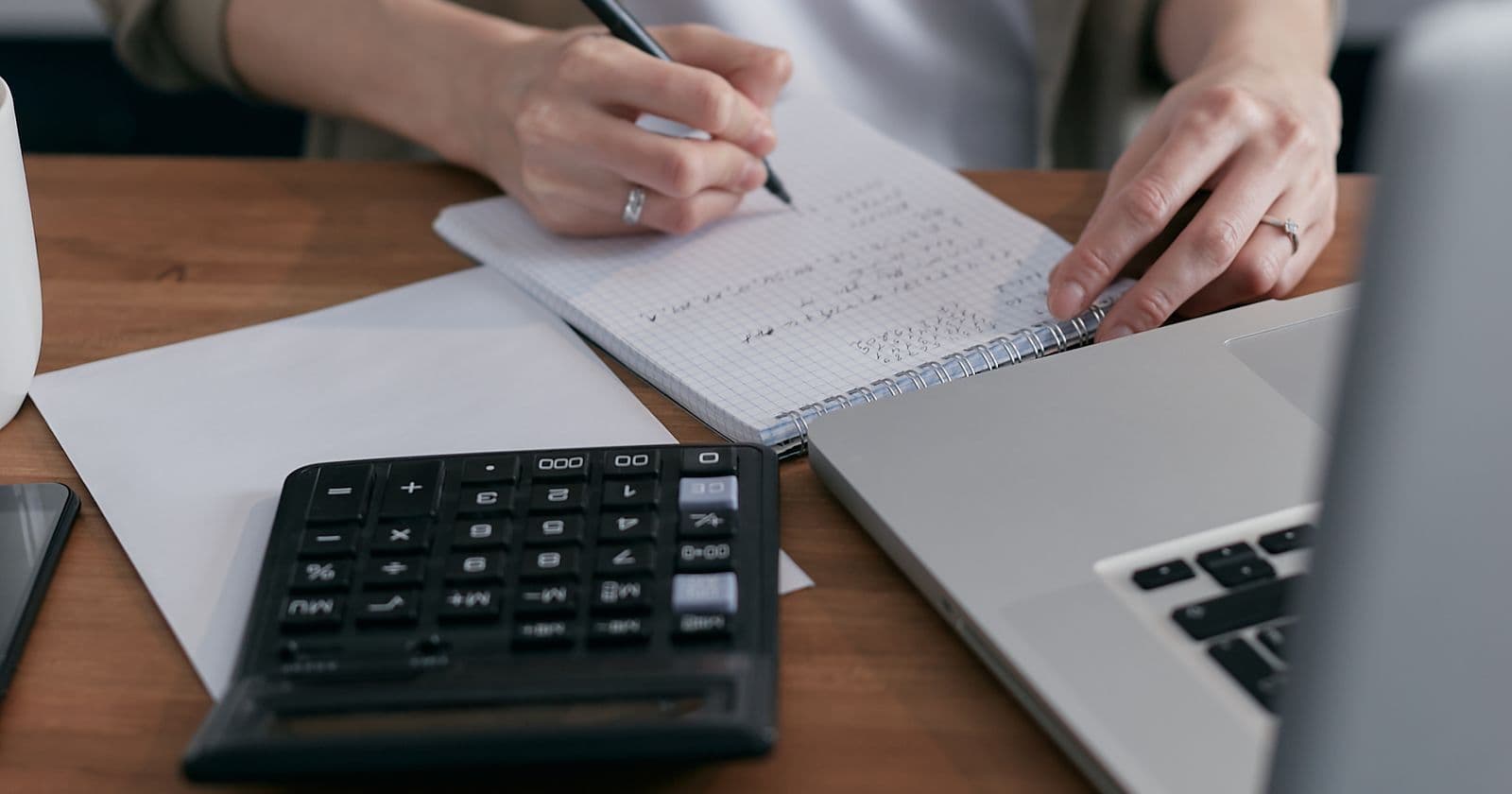 A woman works through figures in a notebook with a calculator beside her laptop