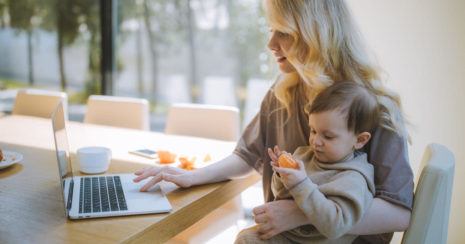A mother works on a laptop at her kitchen table while holding her baby